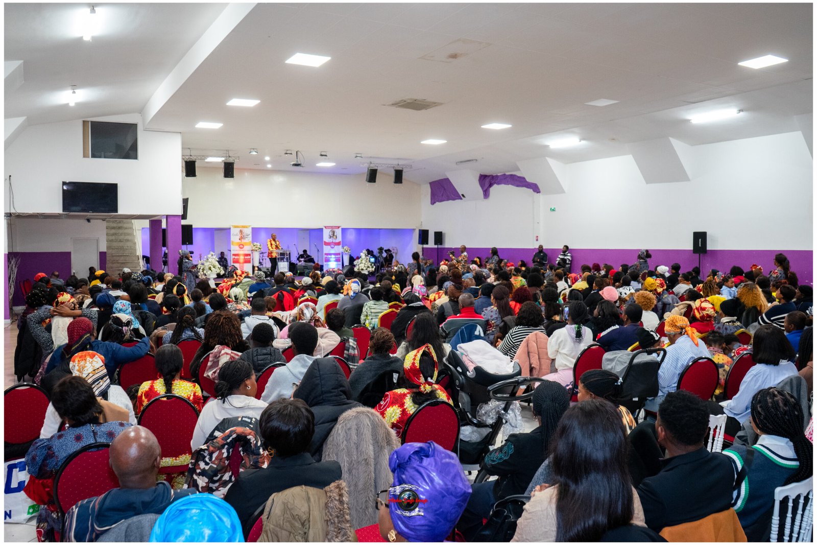 Crowd gathered in a large hall facing a stage with a speaker and banners, purple accents along the walls, many seated attendees wearing colorful clothing.