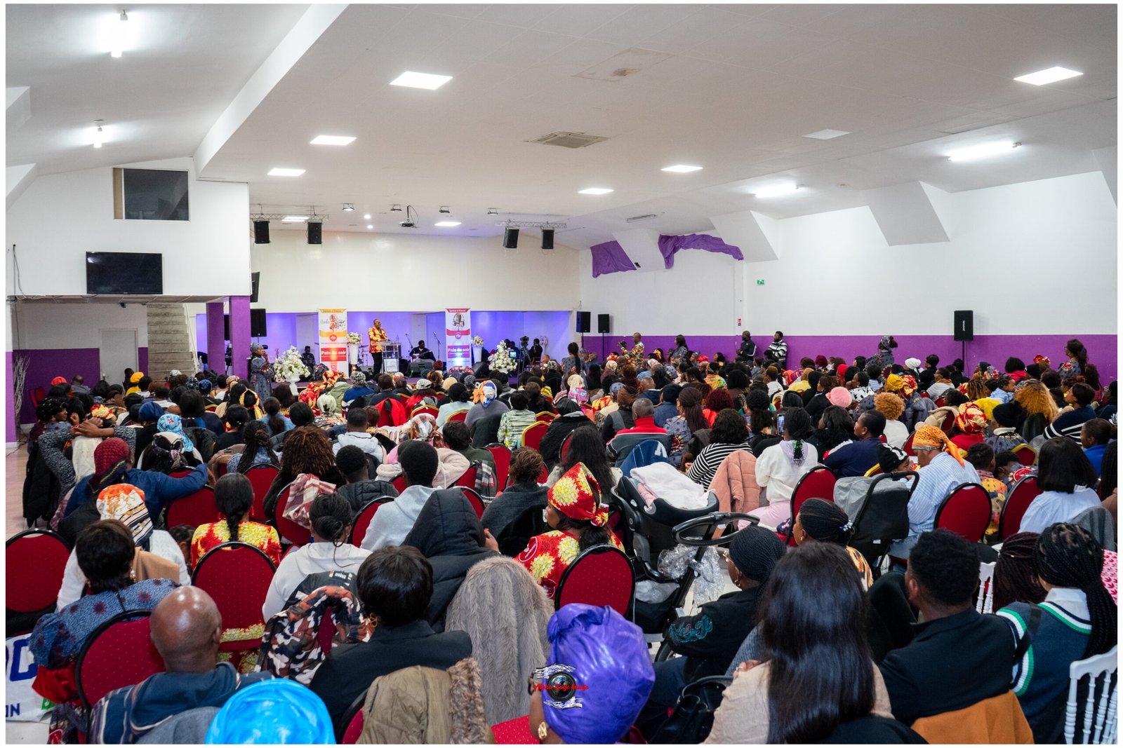 Large crowded hall with people seated in red chairs facing a stage where a speaker stands at a podium, banners and flowers visible, purple accents on walls.
