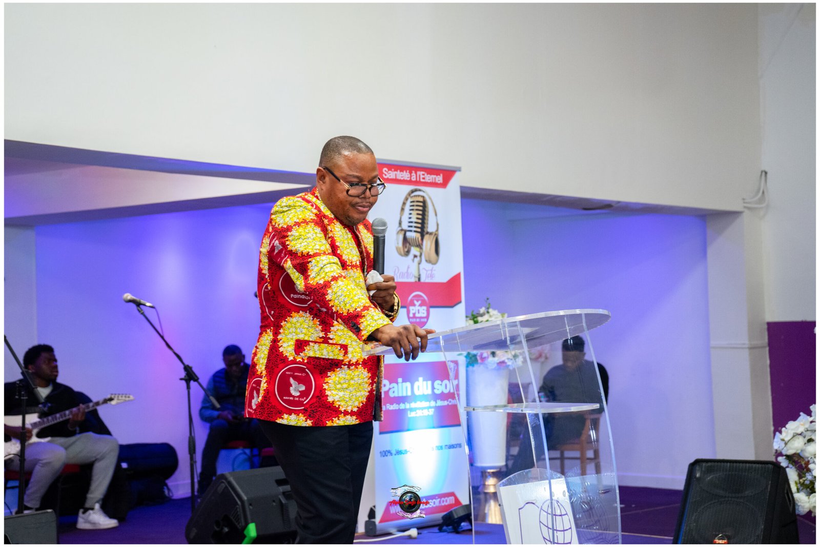 Man in bright African-print shirt speaks into a handheld microphone at a clear lectern on a stage with a blue-lit backdrop.