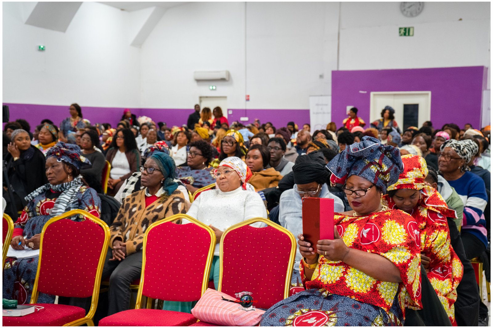 Large crowd of women in colorful head wraps seated in a hall with purple walls at a conference or event. Front row red chairs visible in foreground.