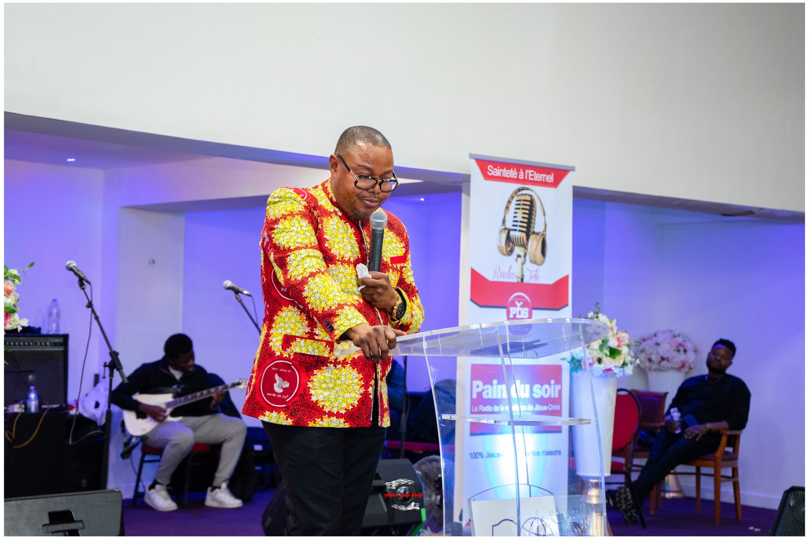 Man in a bright red-and-yellow patterned shirt speaks into a microphone at a clear lectern, with a band and banners behind him.
