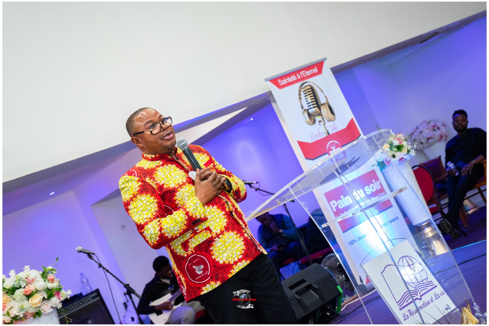 Speaker in a bright red-yellow patterned shirt speaks into a microphone at a transparent podium on a stage with banners and flower arrangements behind him.