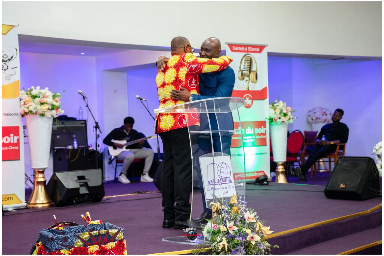 Two men embrace on a stage podium during a ceremony, with a guitarist and banners in the background.