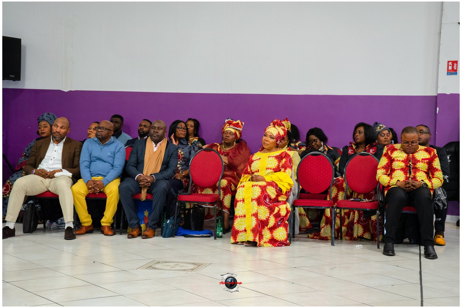 Group of people seated in chairs along a purple wall at a cultural event, wearing colorful traditional outfits and suits.