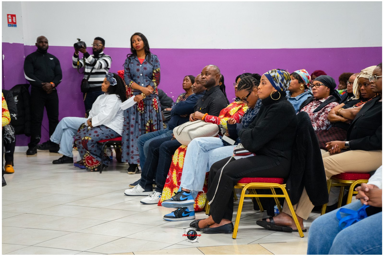 Audience seated in a hall with a purple wall; attendees watch an event, some in colorful traditional clothing, a photographer stands left with a camera.