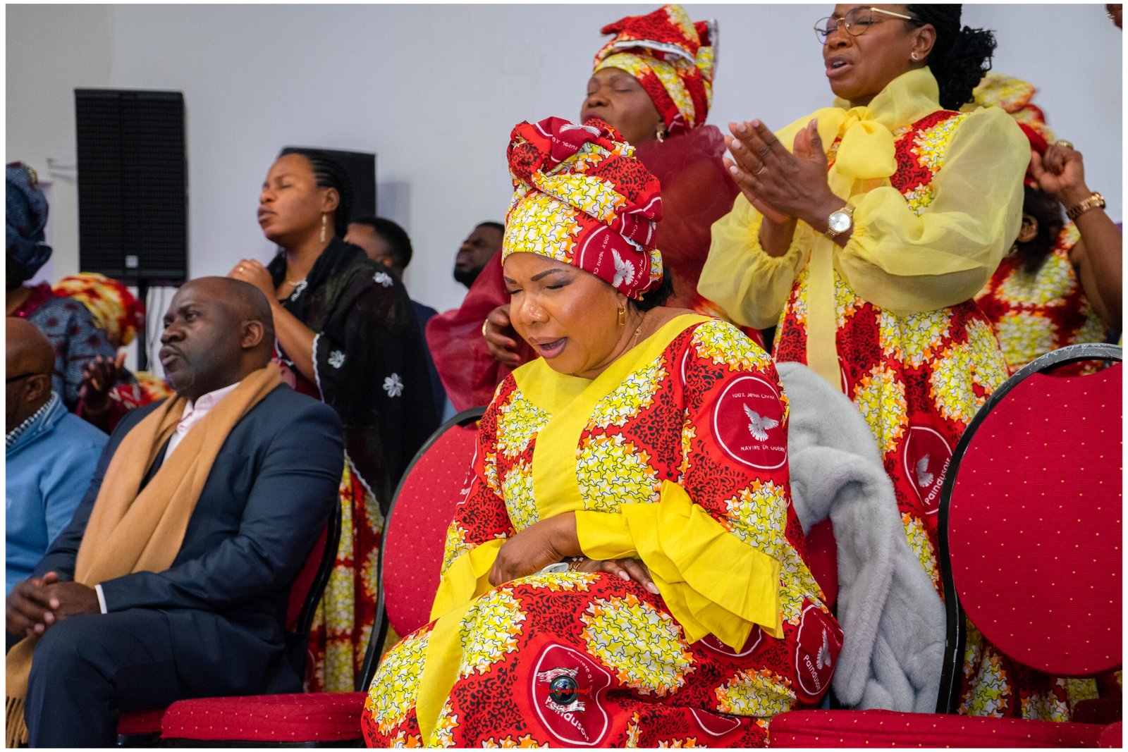 Woman in yellow traditional dress and headwrap, eyes closed, seated among others clapping at a cultural event.