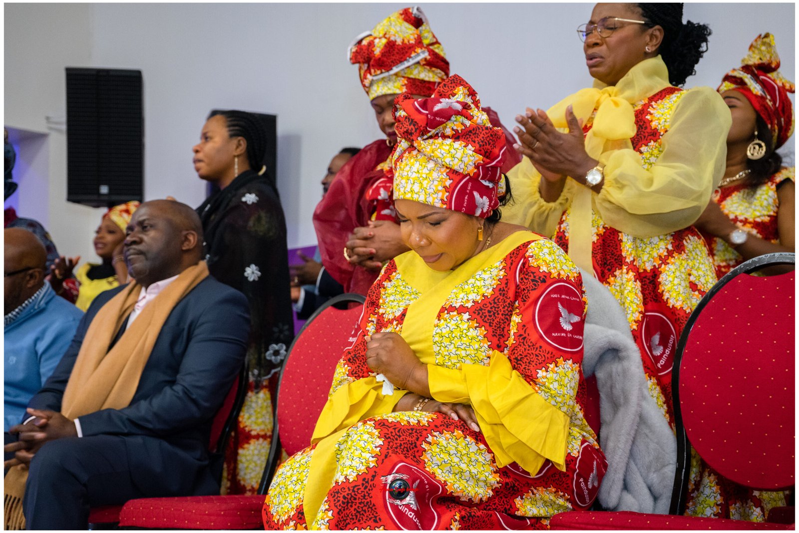 Woman in a yellow and red patterned dress with a matching headwrap, praying and seated among others at a cultural event.