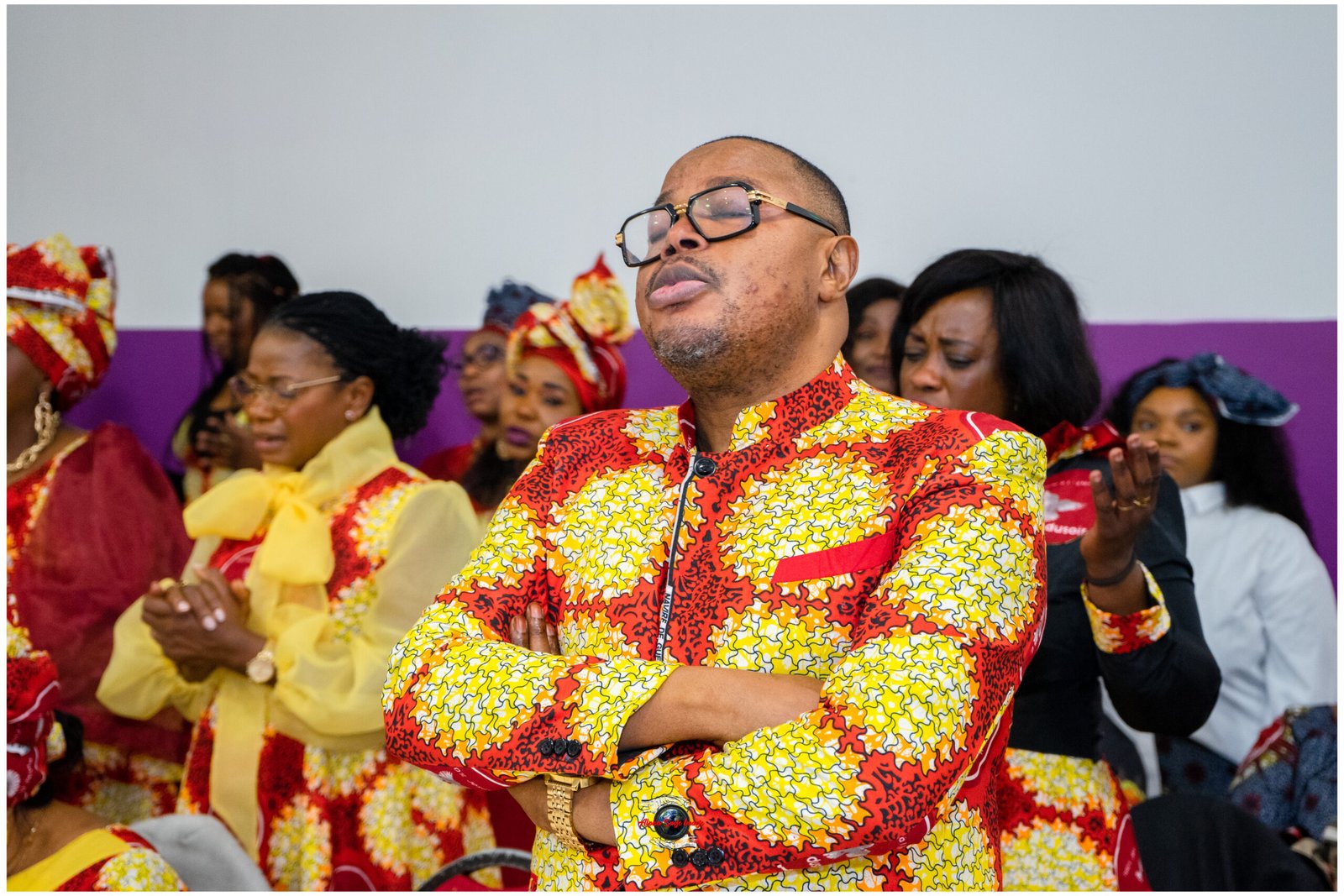 Man in a bright red-yellow African print shirt dancing with arms crossed, wearing glasses, at a celebration.
