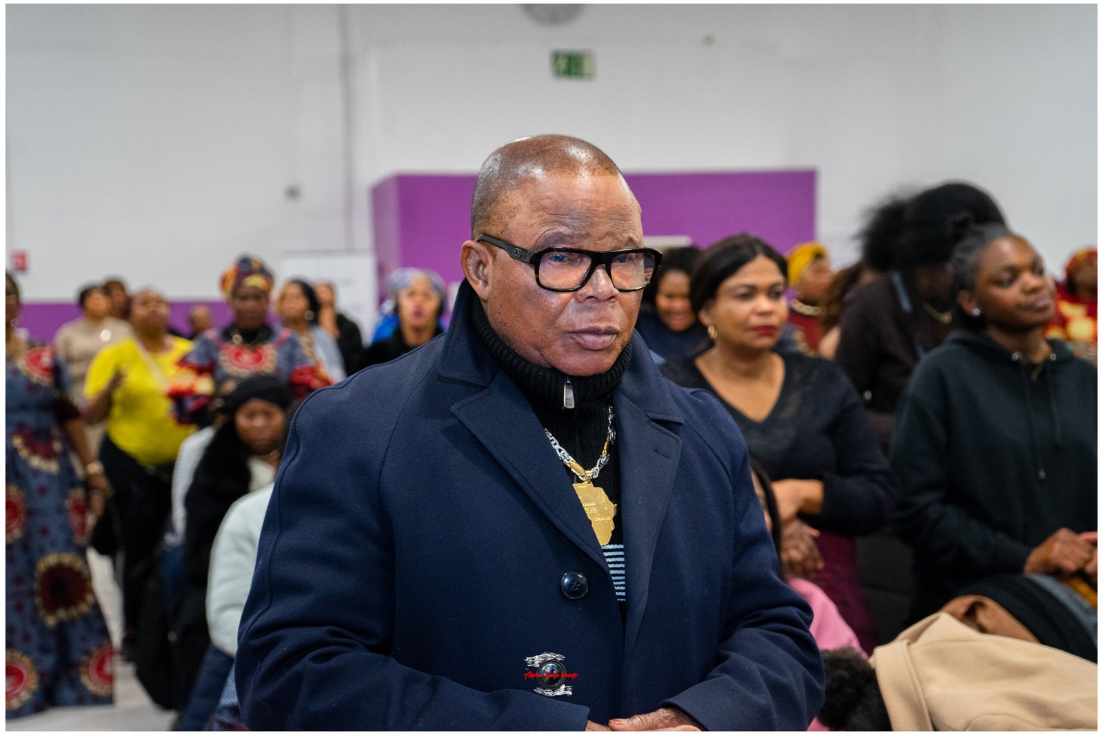 Man wearing glasses and a dark blue coat with a gold pendant, standing at a crowded indoor event foregrounded by attendees in the background.