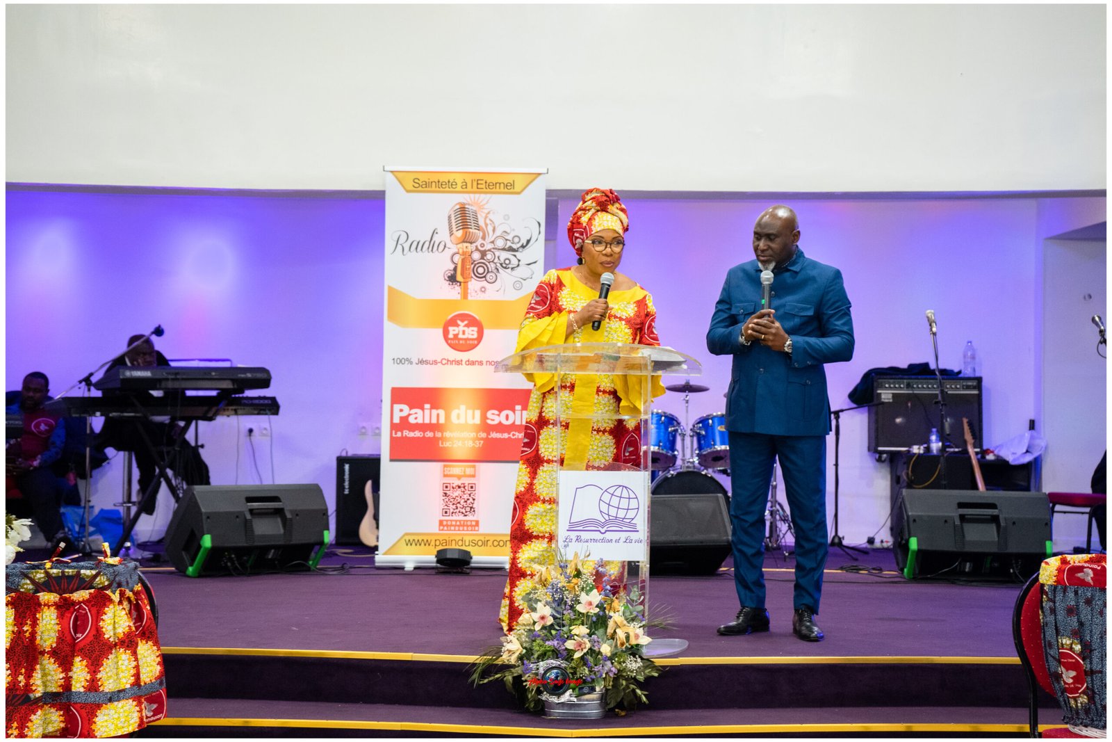 Woman in a bright traditional dress and headwrap speaks at a podium beside a man in a blue suit during a radio event on stage.