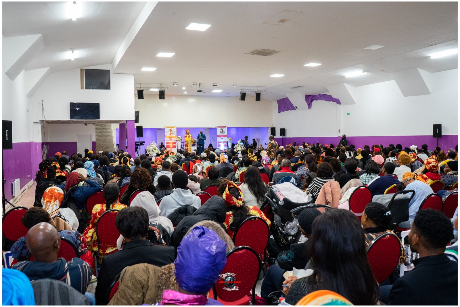 Crowded indoor hall with rows of red chairs filled by people in colorful clothing watching a stage performance with banners and speakers at the front.