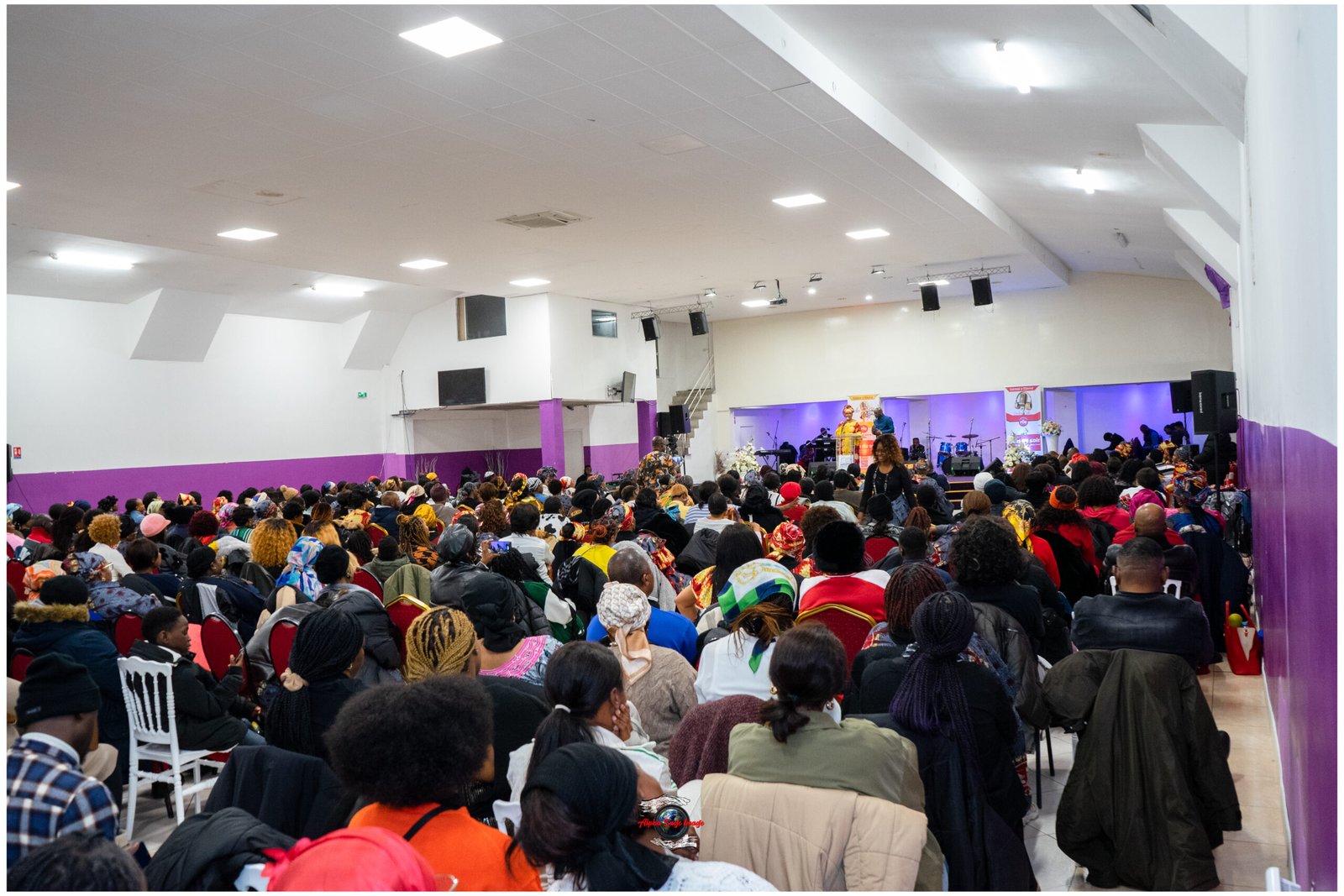 Crowd seated in a large indoor venue watching a stage performance with musicians, purple-walled room in the background.