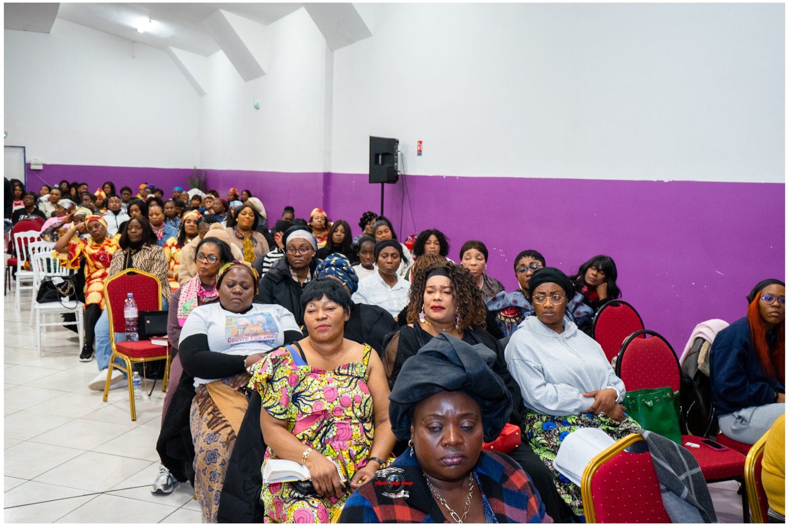 Large group of diverse people seated in rows at an indoor event, facing the camera, with a two-tone white and purple wall behind them.