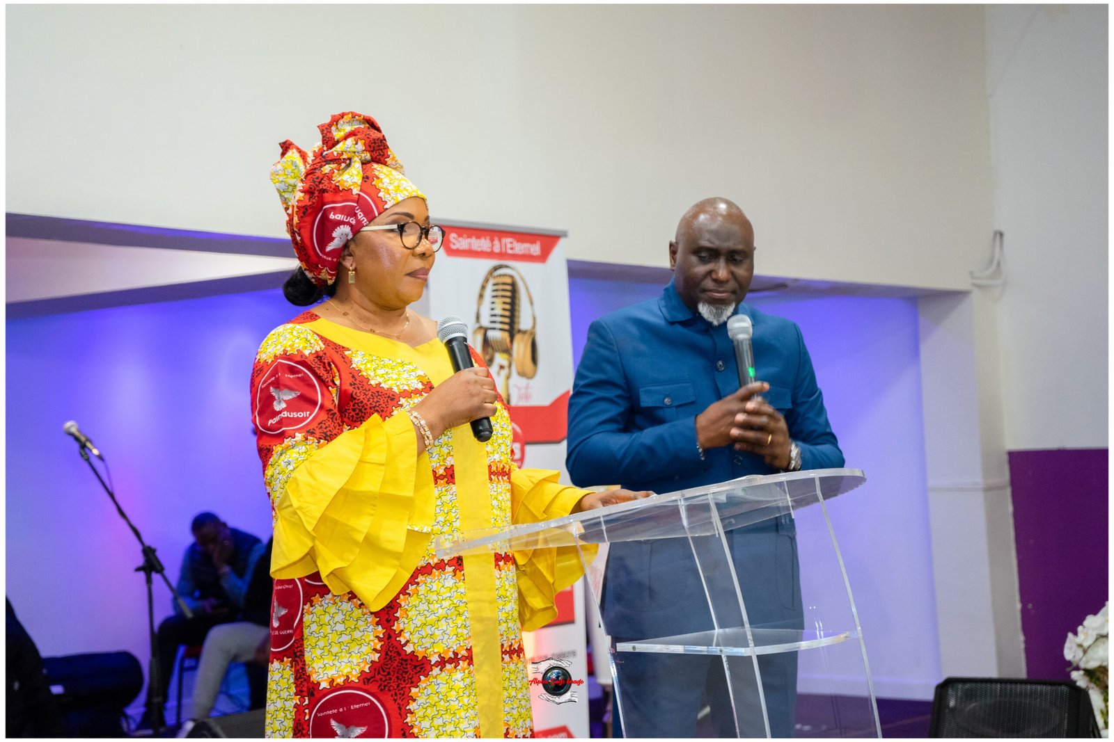 Woman in a colorful African print dress and head wrap speaks into a microphone at a clear podium, with a man in a blue suit beside her on stage.