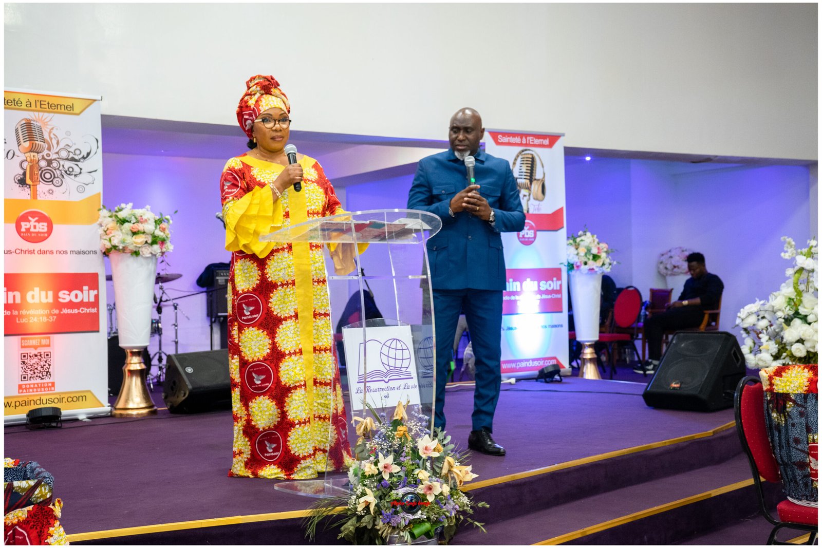 Woman in a bright red and yellow African print dress speaks at a clear podium with a microphone on a decorated stage, a man in a blue suit stands nearby.