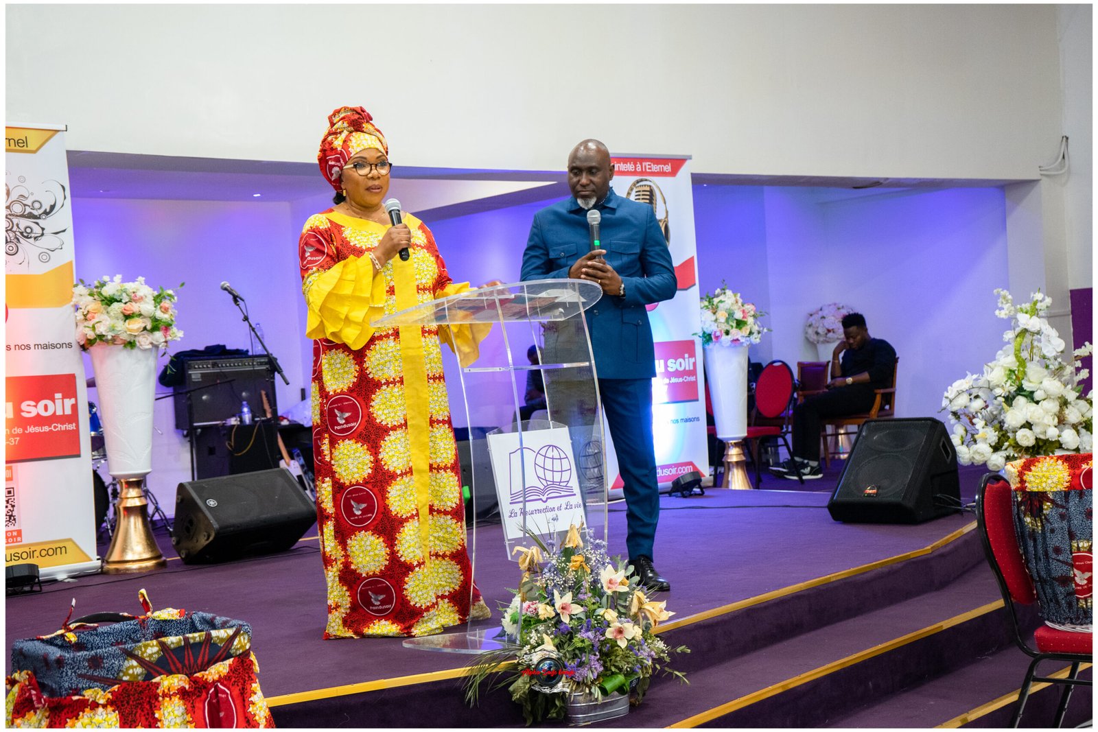 Woman in bright African attire and headwrap speaks into a microphone at a podium on a decorated stage, a man in a blue suit stands beside her.
