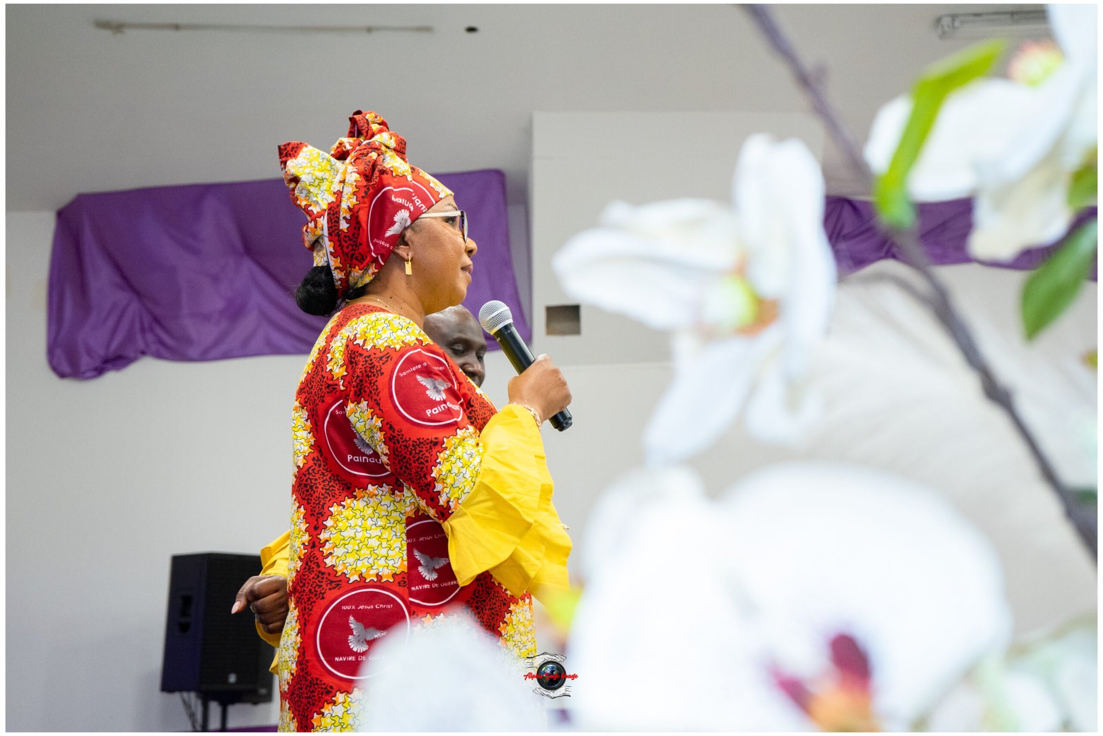 Woman in bright red-yellow patterned outfit and headwrap speaks into a microphone at a stage event, with others blurred in foreground.