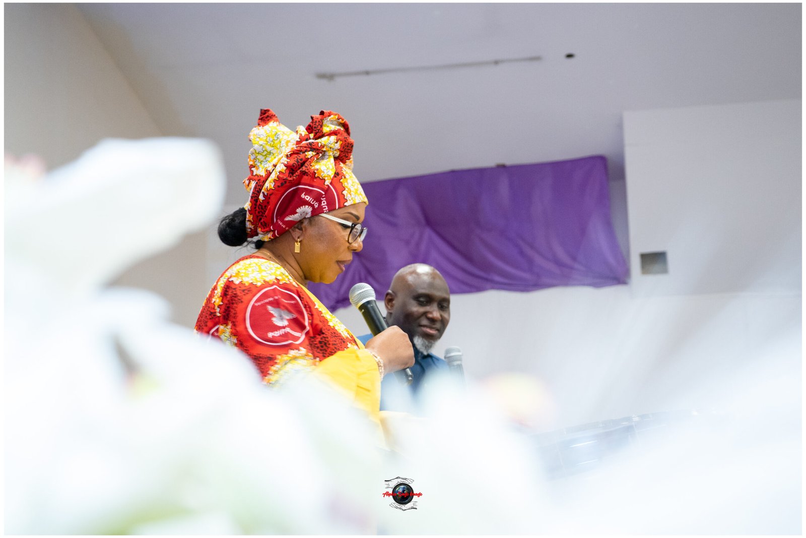 Woman in a bright red and yellow headwrap and dress speaks into a microphone at an indoor event, with a man in the background.