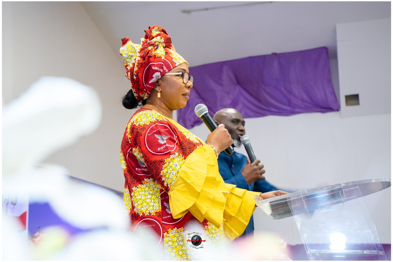 Woman in a bright red and yellow patterned dress and headwrap speaks into a handheld microphone at a podium, with a man in blue behind her.