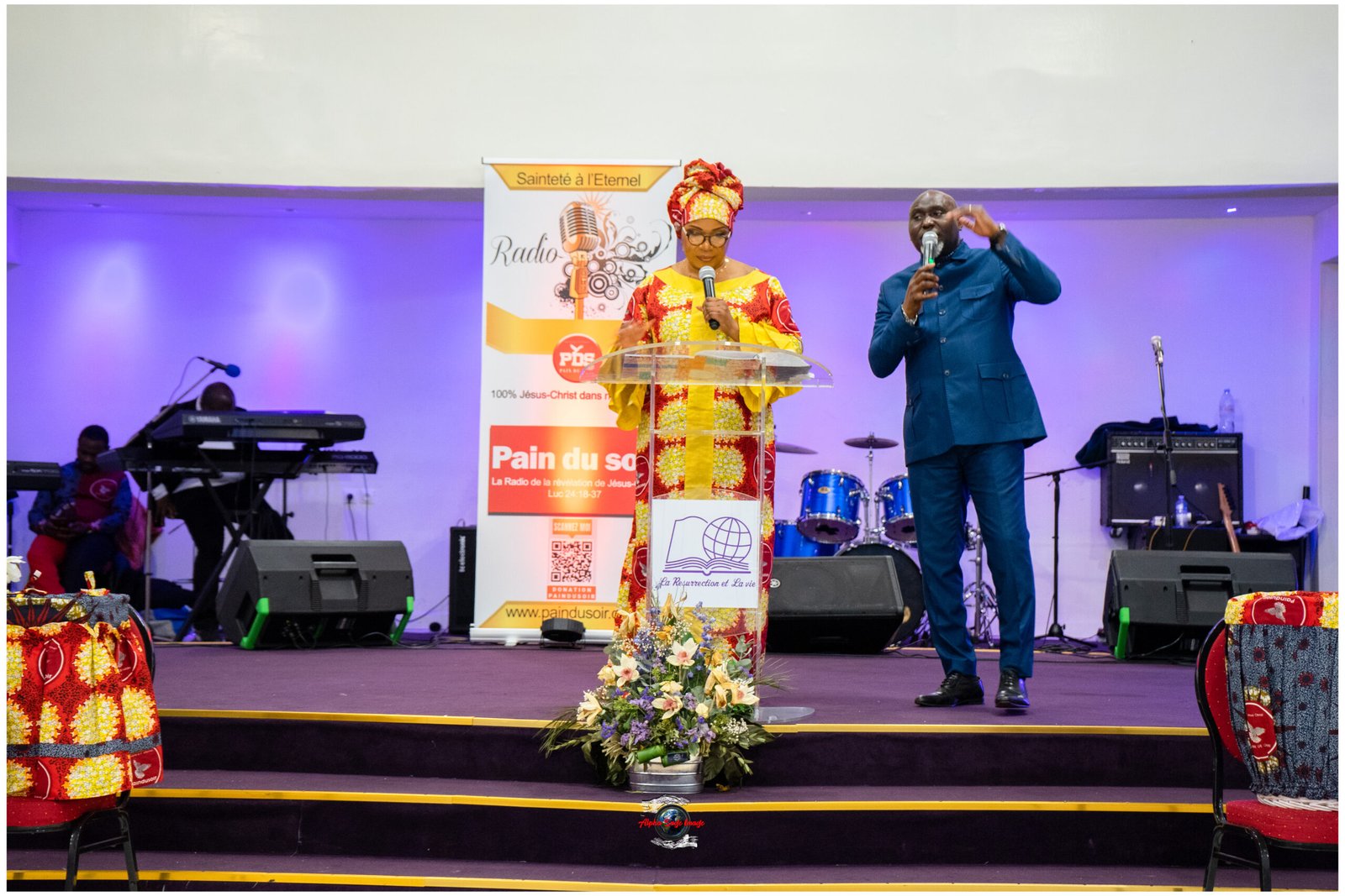 Woman in bright, patterned traditional dress and headwrap speaks at a clear podium on a stage, a man beside her holding a microphone; instruments in the background.