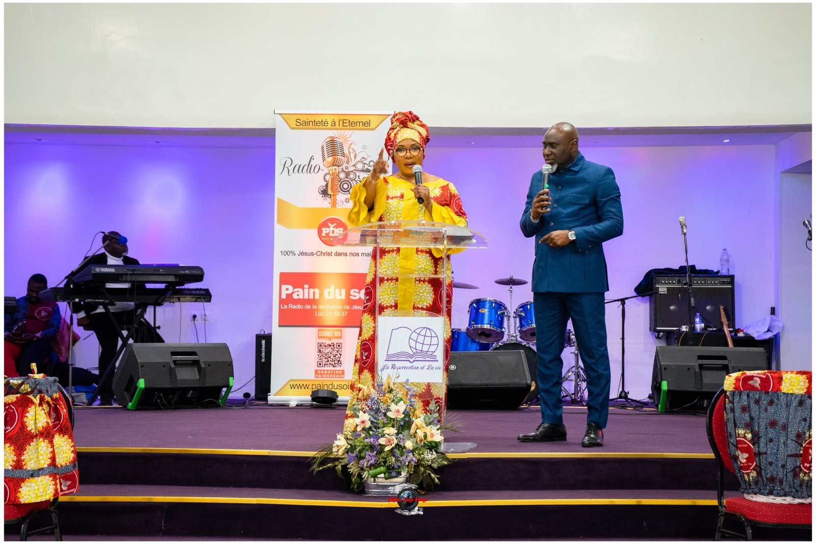 Two presenters stand on a stage speaking into microphones; the woman wears a bright yellow and red patterned dress with a matching headwrap, and the man wears a blue suit. A banner behind them suggests a radio event, with instruments and equipment visible on stage.