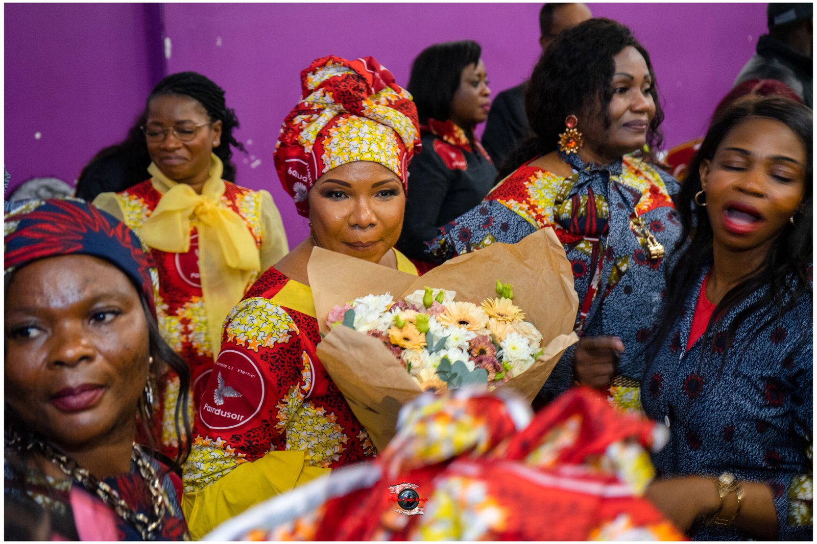 Group of women in vibrant patterned outfits, one in center holds a bouquet of flowers at a celebration with a purple backdrop.
