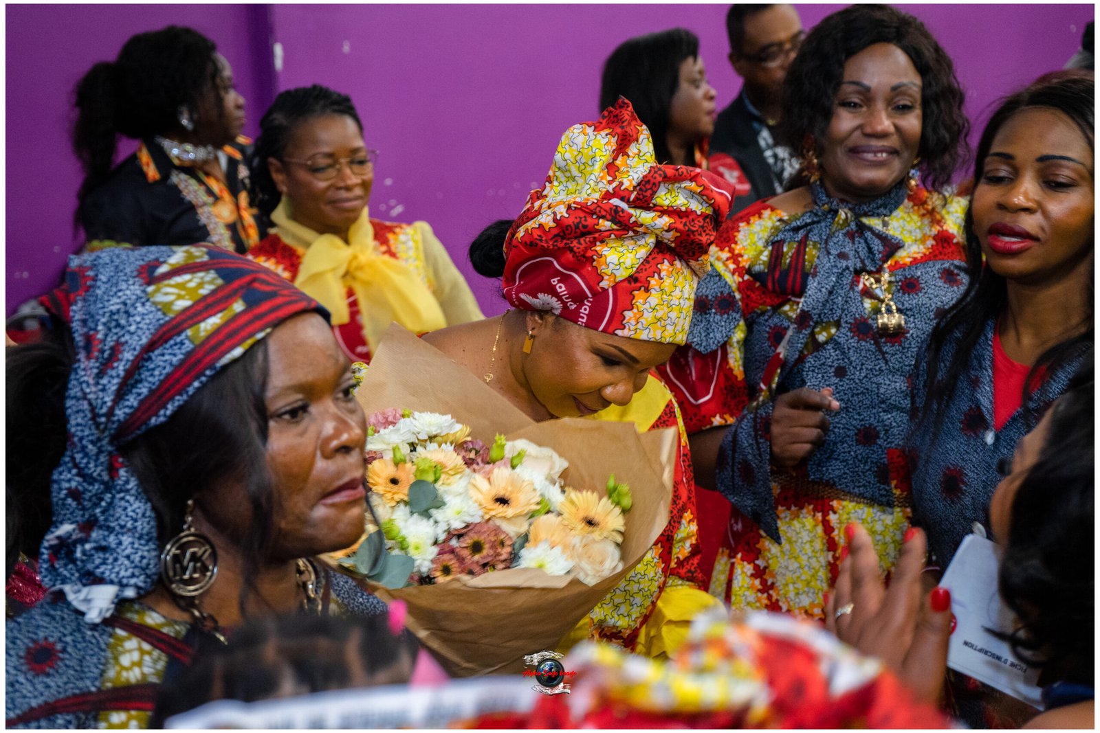 Group of women in bright African print dresses at a celebration, one woman holding a bouquet of flowers.