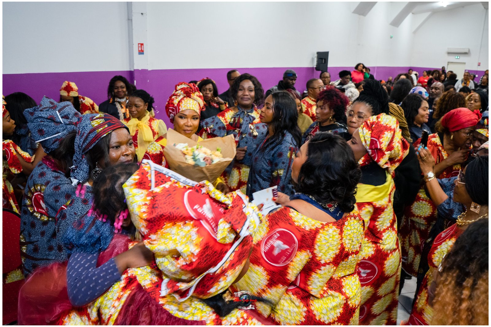 Large crowd of people in vibrant matching Ankara prints at an indoor celebration, center holding a bouquet.