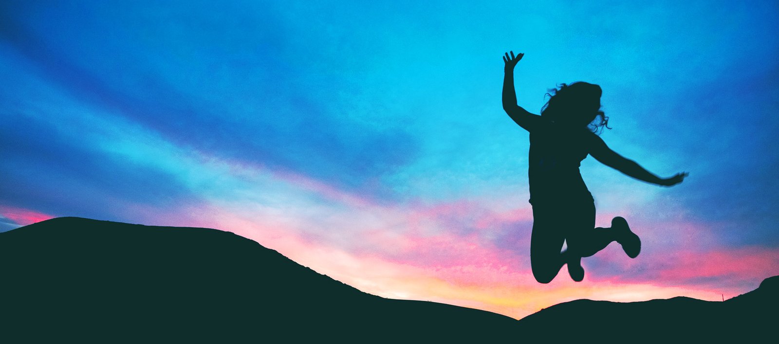 Silhouette of a person jumping with arms raised on a hill at sunset, against a blue-pink sky.