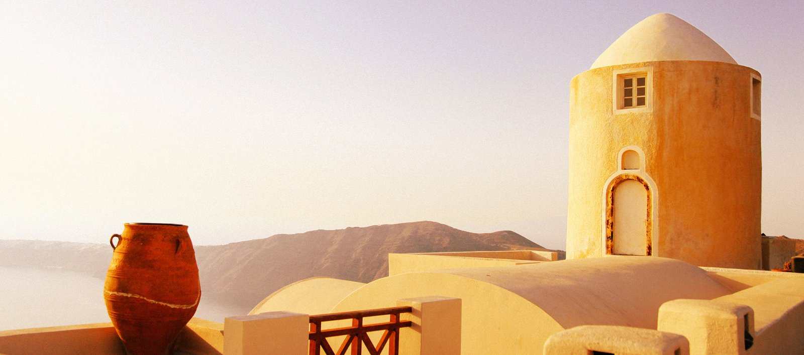 Sunlit white-washed domed building on a terrace, with a large terracotta pot in the foreground and distant hills behind, during golden hour.