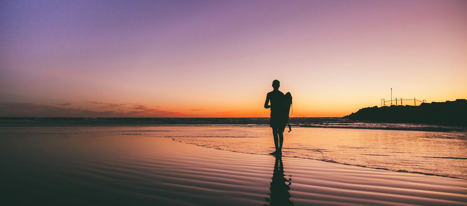 Silhouette of a person carrying a surfboard along the beach at sunset, with an orange-pink sky and calm water reflection.