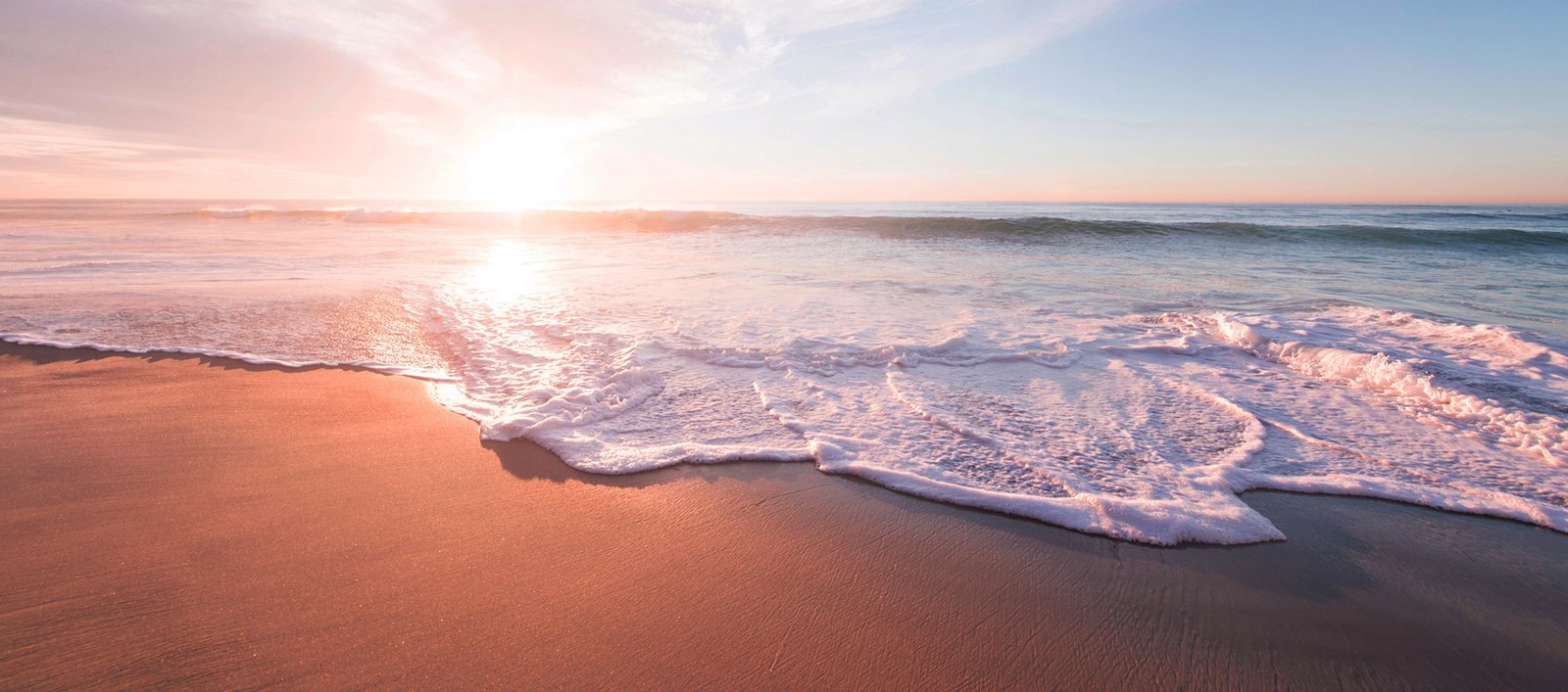 Sunrise over a sandy beach with soft pink clouds and gentle waves washing the shore.
